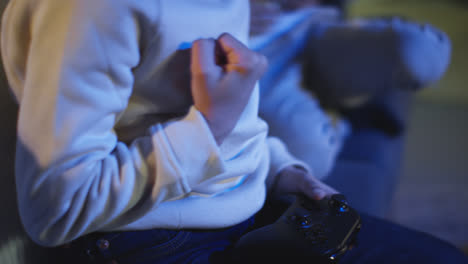 Primer-Plano-De-Un-Niño-En-Casa-Celebrando-Jugar-Con-Una-Consola-De-Juegos-De-Computadora-En-La-Televisión-Sosteniendo-Controladores-A-Altas-Horas-De-La-Noche